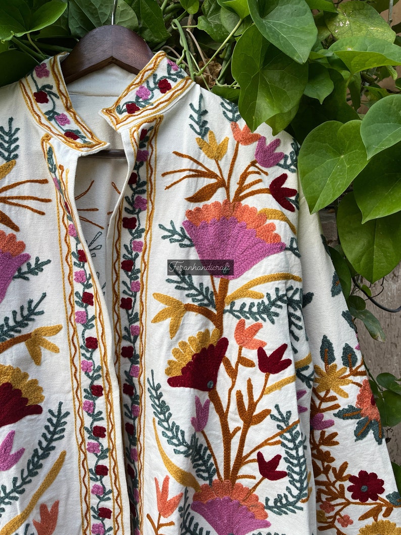 May include: A white cotton jacket with colorful floral embroidery. The embroidery features pink, red, orange, and yellow flowers with green leaves. The jacket has a mandarin collar and a front closure.