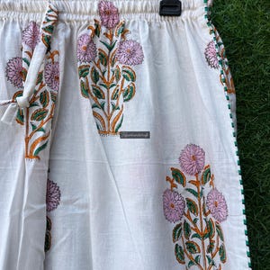 May include: White cotton pants with a floral print in shades of pink, green, and orange. The pants have an elastic waistband and a drawstring tie. The pants have a green trim along the sides.