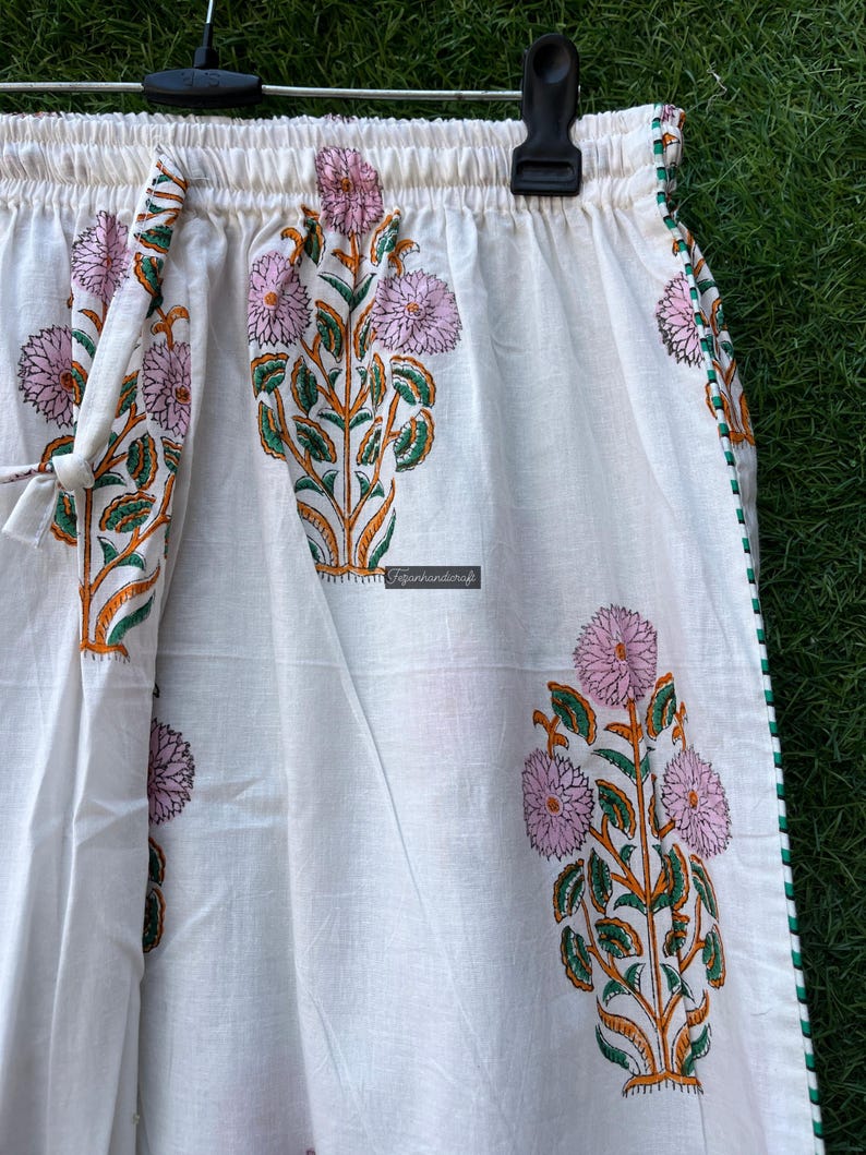 May include: White cotton pants with a floral print in shades of pink, green, and orange. The pants have an elastic waistband and a drawstring tie. The pants have a green trim along the sides.