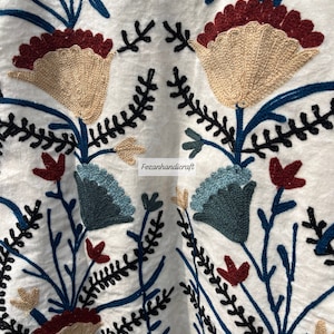 May include: White fabric with embroidered floral design in shades of blue, red, and beige. The embroidery features intricate details and a delicate pattern.