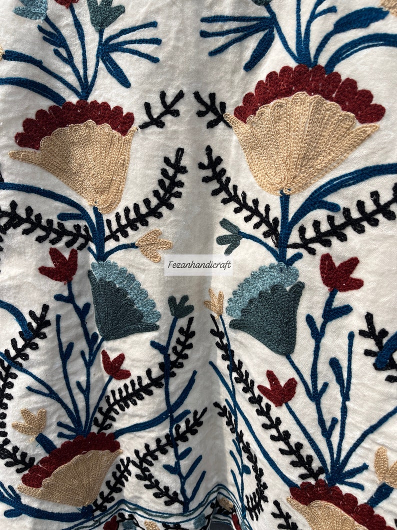 May include: White fabric with embroidered floral design in shades of blue, red, and beige. The embroidery features intricate details and a delicate pattern.