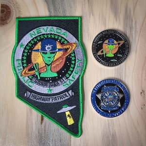 May include: A green, black, and white embroidered patch with the text "Nevada Highway Patrol" and a green alien with a peace sign. The patch is surrounded by stars and a ring. Two silver coins with alien and police themes.