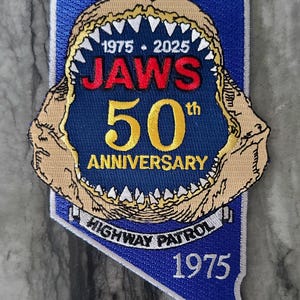 Op de afbeelding: Geborduurde patch met een open haaienbek en de tekst "JAWS 50th ANNIVERSARY" in rood en goud. De blauwe patch heeft de vorm van Nevada en de tekst "HIGHWAY PATROL 1975".