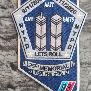 May include: A shield-shaped embroidered patch in blue, white, and grey. It features the Twin Towers, the text 9/11/2001, 9/11/2026, AA77, AA11, UA175, NYFD, LETS ROLL, 25TH MEMORIAL FOR THE 2976, and a 93 logo with red, white, and blue stripes.