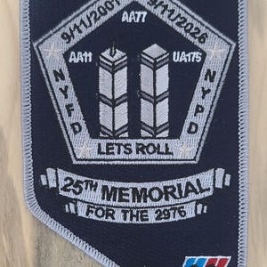 May include: A navy blue patch with white embroidered text and details. The patch commemorates the 25th memorial of the 9/11 attacks, featuring the Twin Towers, the dates 9/11/2001 and 9/11/2026, and the phrase "Let's Roll."