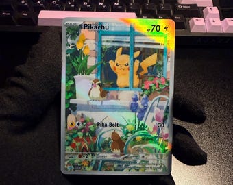 Può includere: Una carta collezionabile olografica di Pikachu che mostra il personaggio che guarda fuori da una finestra con un uccellino marrone sul davanzale. La carta riporta il testo Pikachu, 70 HP, Pika Bolt e 30 danni, su uno sfondo di giardino fiorito.