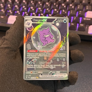 Peut inclure: Une carte à collectionner Gengar ex avec 350 PV, représentant un Gengar violet dans une bulle sur un fond de cimetière. La carte affiche la capacité Gnawing Curse et l'attaque Tricky Steps, avec du texte détaillant les règles du jeu.
