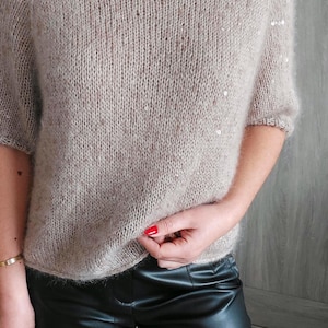Può includere: Un maglione beige a maniche corte con scollo tondo e sottili paillettes sulle maniche. Il maglione è realizzato in morbido tessuto a maglia strutturato. La persona indossa pantaloni di pelle neri.