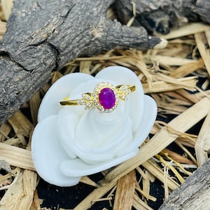 Puede incluir: Un anillo de oro con una gran piedra preciosa ovalada de color púrpura rodeada de piedras preciosas más pequeñas y transparentes.