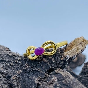 May include: A gold ring with a purple gemstone in the centre, shaped like a bow. The ring is resting on a piece of weathered wood.