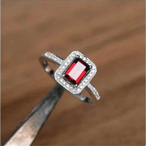 Puede incluir: Un anillo de plata con una piedra preciosa roja engastada en un halo de piedras blancas más pequeñas.