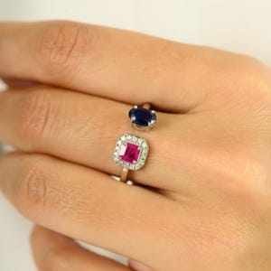 Puede incluir: Un anillo de dos piedras con una gema ovalada azul oscuro y una gema rosa cuadrada rodeada de pequeños diamantes. El anillo está engarzado en una banda plateada, mostrando un diseño moderno y abierto. El anillo se muestra en la mano de una persona.