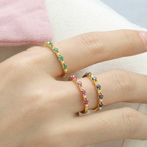 May include: Three gold rings with different coloured gemstones. The top ring has green gemstones, the middle ring has red gemstones, and the bottom ring has blue gemstones.