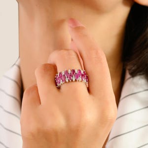 May include: A silver ring with alternating rows of oval-cut, bright pink gemstones and small, clear gemstones. The ring is worn on a finger, highlighting its detailed design and sparkling appearance.