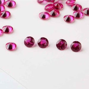 Puede incluir: Una colección de pequeñas piedras preciosas redondas de color rosa. Las piedras preciosas están esparcidas sobre una superficie blanca.