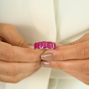 May include: A silver ring with five rectangular, bright pink gemstones. The ring is held between two hands, highlighting the polished stones and the ring's design. The gemstones are set closely together, forming a continuous band of colour.