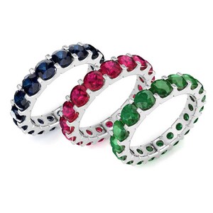 May include: Three eternity bands set with gemstones. The bands are made of white metal and feature a row of round, bezel-set gemstones. The gemstones are blue, red, and green.