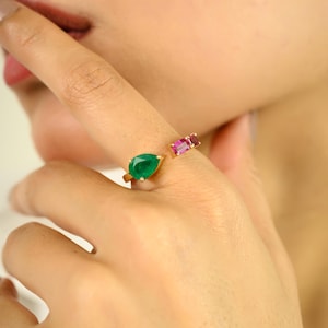 Peut inclure: Une bague en or ornée d'une pierre verte en forme de poire et de deux pierres roses rectangulaires. La bague ouverte présente les pierres précieuses de chaque côté du doigt. La bague est portée sur le doigt d'une personne.