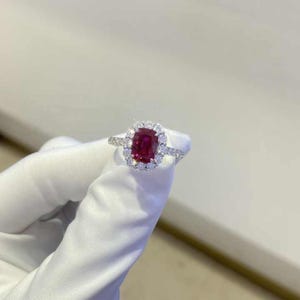 以下が含まれることがあります： 赤い宝石が、小さな白い宝石のハローで囲まれたリング。リングは白い金属でセットされています。