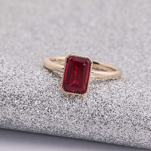 Puede incluir: Un anillo de oro con una gran piedra preciosa roja rectangular. La piedra preciosa está engastada en un engaste de bisel de oro con un borde decorativo.