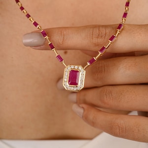 May include: A gold necklace featuring a large, rectangular ruby pendant surrounded by small diamonds. The necklace chain is composed of small, rectangular rubies set in gold. The jewelry is held by a hand, showcasing the necklace's design and color.