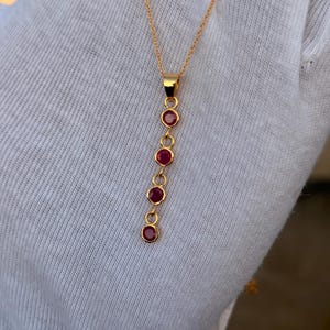 May include: A gold chain necklace featuring a vertical drop pendant composed of four round, bezel-set red gemstones arranged in a linear sequence. The pendant is suspended from a delicate gold link chain, showcasing a polished metal finish.