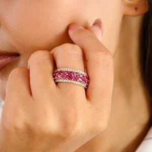 May include: A gold ring with two rows of pink gemstones, possibly rubies or pink sapphires, separated by a row of smaller, clear stones. The ring is displayed on a finger, highlighting the design and colour of the jewellery.