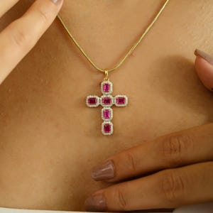 Pode incluir: Um colar dourado com um pendente de cruz. A cruz é adornada com pedras retangulares de cor rubi e rodeada por pequenas pedras transparentes. O colar é exibido sobre um fundo cor de pele.