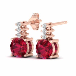 May include: A pair of rose gold earrings with a round red ruby gemstone and three small round diamonds. The earrings have a simple design with a single ruby and three diamonds.