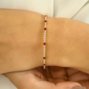 May include: A delicate gold bracelet featuring alternating red gemstones and clear crystals. The bracelet is a slender design, showcasing a line of vibrant red stones interspersed with smaller, sparkling crystals. The bracelet is worn on a wrist.