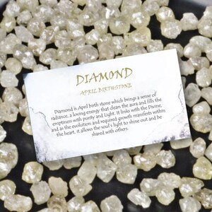 May include: A collection of rough, uncut diamonds with a card that reads "DIAMOND APRIL BIRTHSTONE". The diamonds are a pale yellow color and are scattered across a dark surface. The card provides information about the April birthstone.