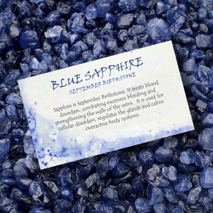 May include: A close-up of blue sapphire gemstones with a card that reads "BLUE SAPPHIRE SEPTEMBER BIRTHSTONE". The card describes the gemstone's properties. The gemstones are a deep blue color and have a rough texture.