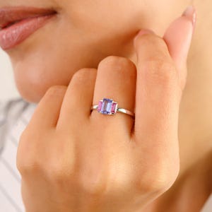 May include: A silver ring featuring a rectangular, light blue gemstone flanked by two smaller, rectangular pink gemstones. The ring is set on a woman's finger, showcasing the jewelry's design and color contrast.