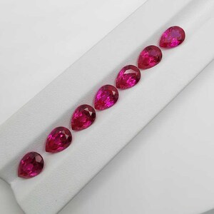 May include: Seven faceted, pear-shaped rubies displayed on a white surface. The gemstones are a vivid, deep pink-red hue. Each gem is cut to reveal internal facets, creating a sparkling effect. The arrangement is diagonal, showcasing the gems' shape.