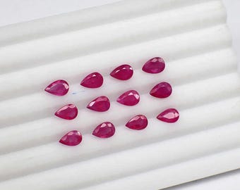 May include: A collection of 12 pear-shaped ruby gemstones, arranged in rows on a white surface. The rubies are a deep red color and have a glossy finish.