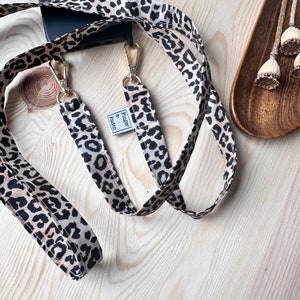 Könnte beinhalten: Ein braunes und schwarzes Handyband mit Leopardenmuster, goldenen Beschlägen und einem kleinen Etikett mit der Aufschrift "Finger & Faden".