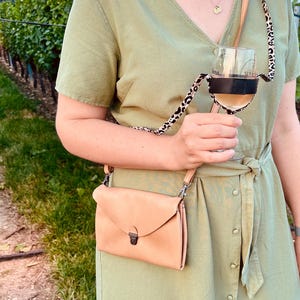 May include: A woman wearing a light green dress with a brown leather crossbody bag and a leopard print wine glass holder. The holder is attached to a glass of white wine.