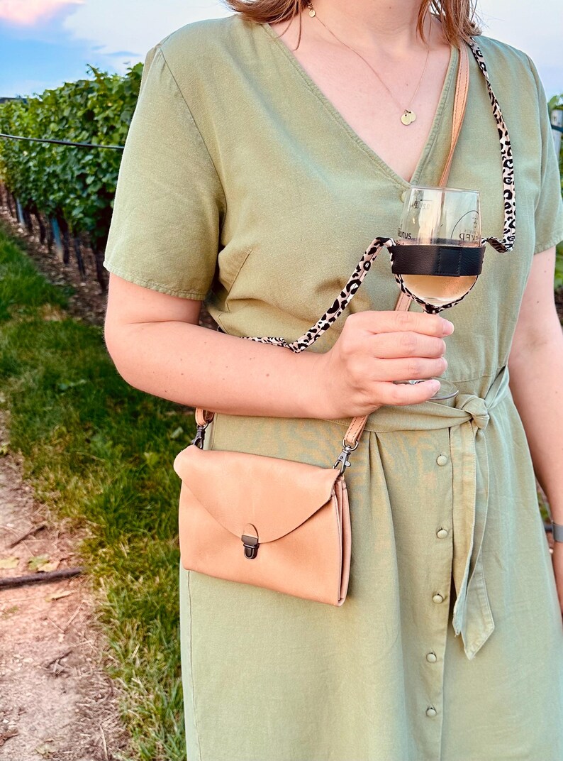 May include: A woman wearing a light green dress with a brown leather crossbody bag and a leopard print wine glass holder. The holder is attached to a glass of white wine.