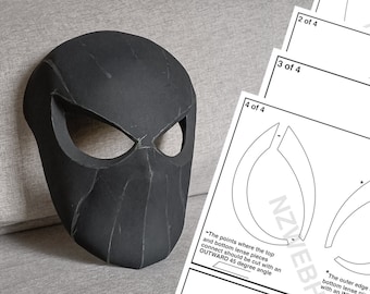May include: A black foam mask with a spider-like design. The mask has two eye holes and a raised, textured design. The mask is sitting on a gray surface. The image also includes a set of instructions for assembling the mask.