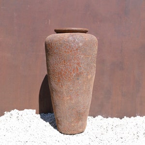 May include: A large, brown, ceramic vase with a textured surface. The vase is standing on a bed of white gravel.