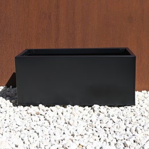 May include: A black rectangular planter sits on a bed of white gravel. The planter is made of metal and has a simple, modern design.