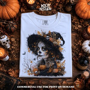 Pu&ograve; includere: Una t-shirt bianca con una stampa grafica di una persona che indossa un cappello da strega e un trucco da teschio, circondata da foglie autunnali, zucche e ragnatele. Il testo "Witchy ArtStudio" &egrave; stampato sopra la grafica sulla maglietta.