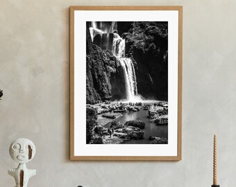 May include: A black and white photograph of a waterfall in a forest setting. The photo is framed in a light wood frame and is displayed on a white wall above a white shelf with various decorative items.