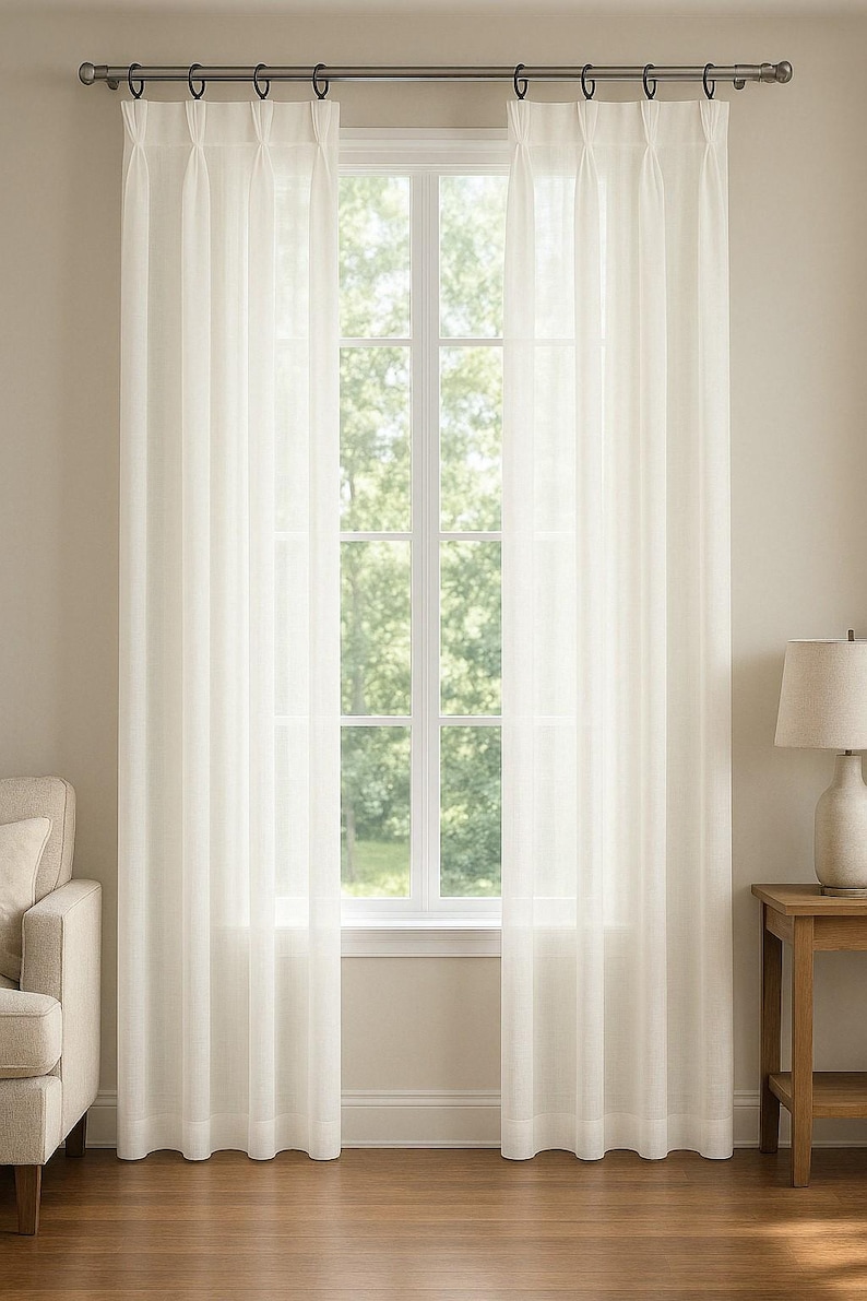 May include: A pair of off-white, semi-sheer curtains featuring a pleated top header. The curtains are suspended from a metallic rod with circular rings, hanging vertically to cover a window that reveals a view of green trees outside.