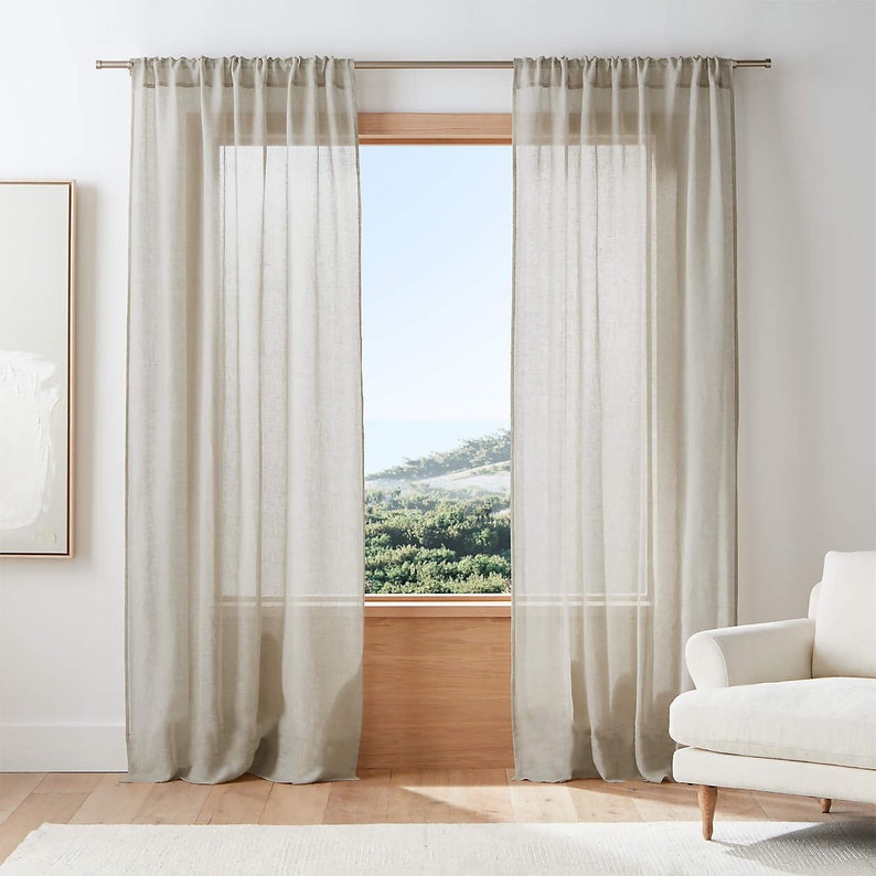 May include: A pair of sheer, light beige linen curtains hang in a window, with a view of a green hillside and blue sky in the background.