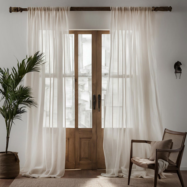 May include: White sheer curtains hang in front of a wooden door with glass panels. The curtains are gathered at the top and hang straight down. The door is closed and has a wooden frame. There is a chair in the foreground.