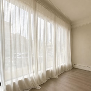 Peut inclure: Rideaux blancs transparents allant du sol au plafond couvrant une grande fenêtre. Le tissu léger et semi-transparent tombe en plis verticaux depuis un système de rail fixé en haut du mur, créant un effet de lumière douce et diffuse.
