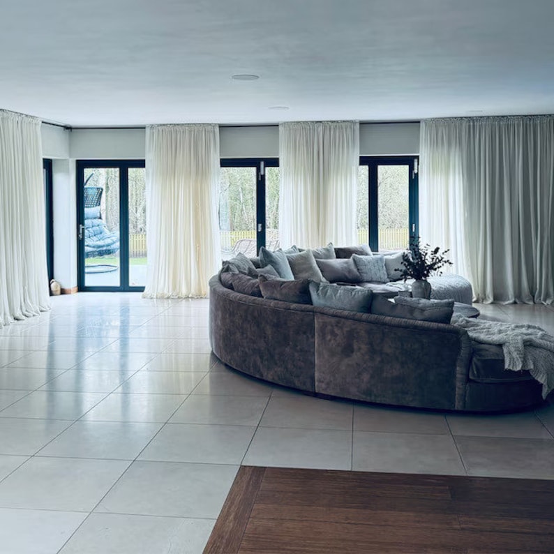 May include: A large, circular, gray sectional sofa with light blue pillows sits in a spacious living room with white curtains and large windows. The floor is tiled in white.