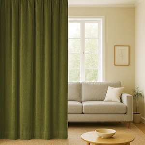 May include: Olive green velvet curtain hanging in front of a window. The curtain is floor-length and has vertical folds. A light grey sofa with a white cushion is visible behind the curtain. A wooden coffee table and a small plant are also in the room.