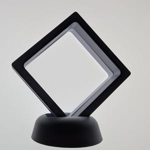 May include: A black floating display case with a square aperture, supported by a rounded base. The case is designed to hold and showcase small objects, with a clear interior space for the item to be displayed.